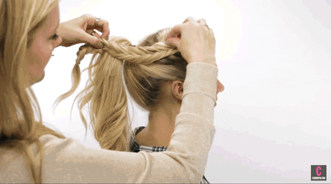 A woman is styling another woman's hair into a braid from the back. the stylist is meticulously weaving the hair strands with focus and precision, in a neutral colored setting.