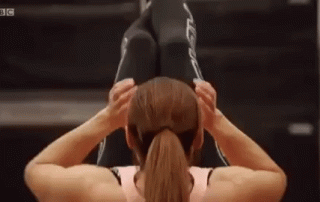 A woman in a gym performs an upside-down sit-up with the assistance of a male trainer, who supports her legs as she exercises.