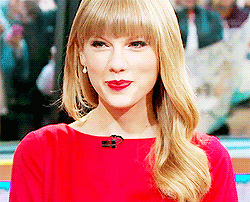 A smiling woman with blonde hair and bangs, wearing a bright red top, sitting in a studio with blurred people visible through a window in the background.