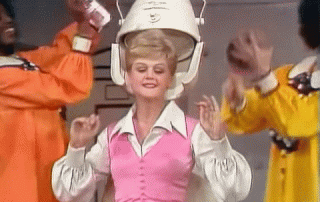 A woman in a pink outfit wears a large, helmet-like hair dryer, dancing joyfully while surrounded by people in colorful costumes clapping to the beat.