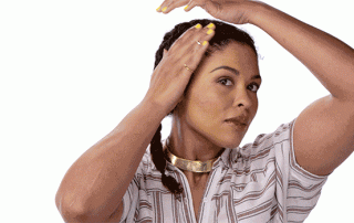 A woman, wearing a striped shirt and a golden choker, making a gesture with her hands around her face as if framing it, looking intently at the camera with a slight smile.