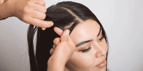 A hairstylist skillfully braids the long, dark hair of a woman, who appears relaxed with closed eyes against a neutral backdrop.