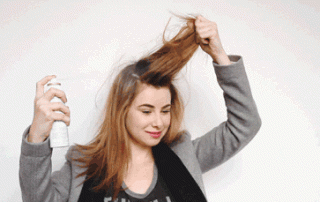 A woman with long hair using hairspray on herself, holding a section of her hair up as she sprays it, smiling, and wearing a gray blazer.