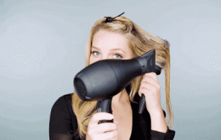 A woman with blonde hair is styling her hair using a black hair dryer and a round brush against a light blue background.