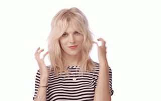 A woman with blonde hair and a striped top gesturing with her hands, looking directly at the camera with a playful expression.