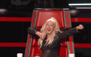 A woman with blonde hair, wearing a black outfit, joyfully hits the red button and throws her arms in the air while sitting in a revolving chair on "the voice".