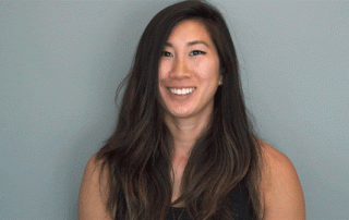 A woman with long dark hair smiling at the camera against a gray background. she is wearing a dark sleeveless top.