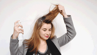 A woman in a gray blazer using hairspray on her lifted, tousled hair against a plain light background. she appears joyful, tending to her hairstyle.