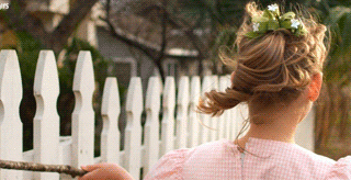 A young girl with a braided updo and a floral crown walks along a white picket fence, holding a stick, in a rural setting.