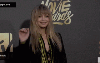 A woman on a red carpet, playfully waving, dressed in a stylish striped suit with her blonde hair styled in bangs, at an awards show with a logo backdrop.