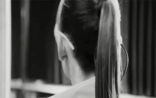 Black and white animated gif showing a woman from the back as she ties her long hair up into a ponytail. a curtain gently sways in the background.