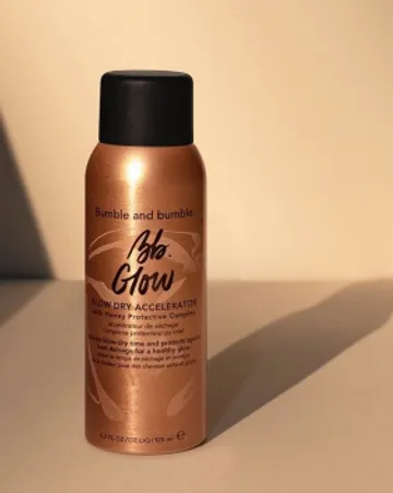 A bottle of bumble and bumble glow blow dry accelerator spray stands against a plain background, illuminated by sunlight that casts a soft shadow. the bottle is metallic bronze with black cap.