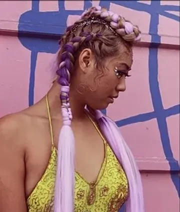 A woman with intricate purple braids and patterns painted on her face, wearing a yellow top, stands against a pink and blue geometric background. she looks downwards in a thoughtful pose.