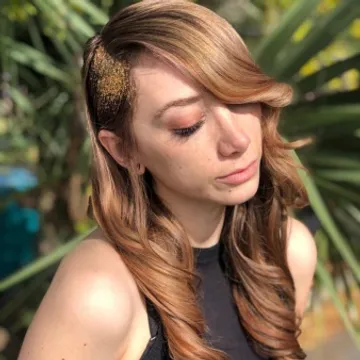 A woman with wavy brown hair adorned with a golden glitter hairpiece, eyes closed, posing in a tropical setting with blurred greenery in the background.