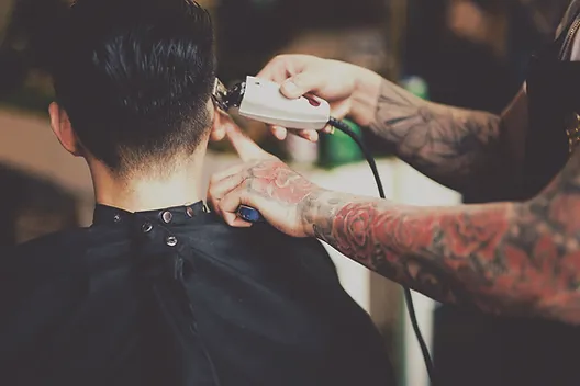 A person with tattooed arms using electric clippers to trim the back of another person's head in a barber shop.