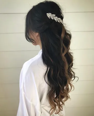 A woman with wavy, dark hair styled in a half-up, half-down hairstyle, secured with a bow-shaped clip with rhinestone embellishments, against a white wooden background.