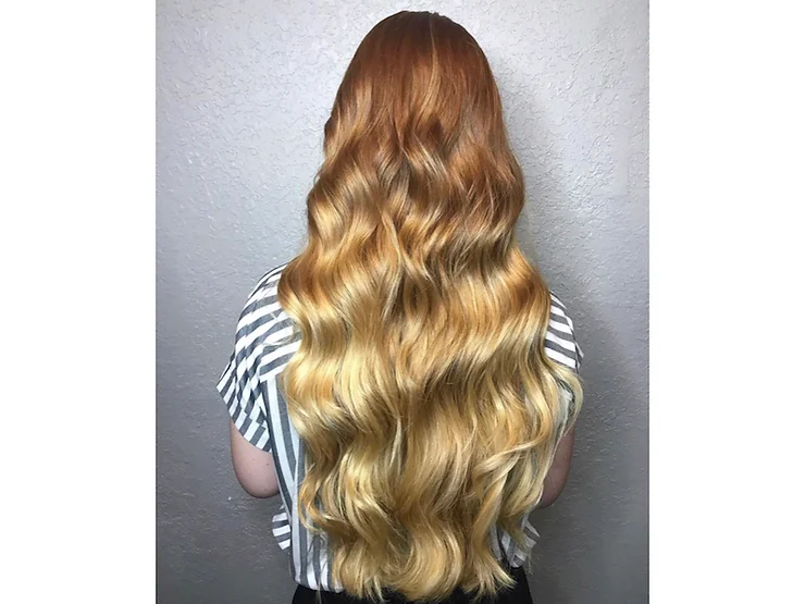 A person from behind showcasing long, wavy hair with a gradient from reddish-brown at the roots to blonde at the ends against a neutral background.