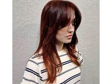 A woman with shoulder-length auburn hair and bangs, wearing a white striped shirt, stands against a textured gray wall, looking to her right.