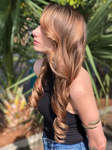 A woman with long, curly blond hair looks away from the camera, standing outdoors with tropical plants in the background. she wears a black tank top and blue jeans.