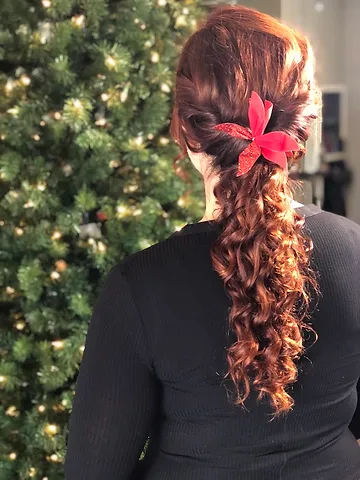 A woman with a curly braid decorated with a red bow sits in front of a twinkling christmas tree.