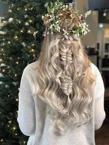 A woman with long, beautifully styled hair adorned with a floral crown stands in front of a christmas tree, showcasing intricate braiding and soft curls.