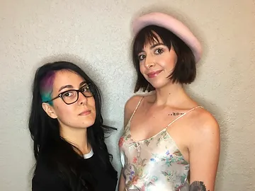 Two women posing together against a beige wall; one with glasses and multi-colored hair, the other wearing a pink hat and floral dress. both are smiling.