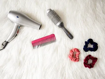 A hairdryer with a round brush, a pink comb, and three colorful scrunchies arranged on a white furry background.