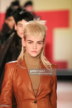 A model with a dramatic blonde mohawk and bold eyebrows is walking the runway, wearing a stylish brown leather blazer over a turtleneck, against a pink and red background.