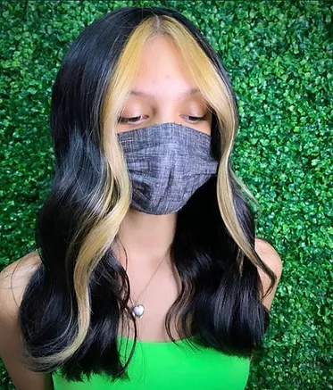 A person wearing a gray mask, with black hair featuring blonde highlights, is standing against a lush green leafy background.