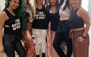 Five women posing together indoors, smiling at the camera. two are wearing playful graphic t-shirts, and the others are in casual chic outfits. the setting has a modern, casual vibe.