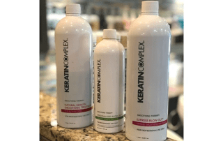 Three bottles of keratin complex hair smoothing therapy products arranged in ascending order by size on a countertop, with the largest on the right.