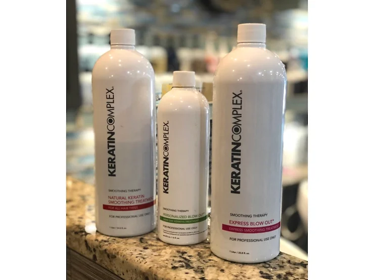 Three bottles of keratin complex smoothing therapy products, varying in size, displayed on a countertop. the largest bottle is on the right, the smallest in the middle.