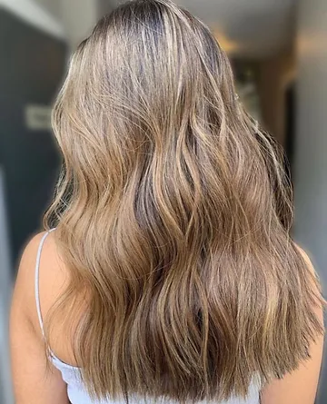 Back view of a person showcasing their long wavy hair with a blend of blonde and brown balayage highlights. the setting appears to be indoors with soft lighting.