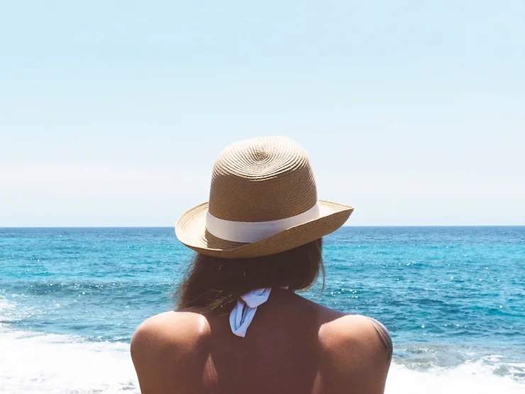 A woman wearing a straw hat and a white ribbon, viewed from behind, looks out over a calm blue sea under a clear sky.