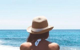 A woman wearing a straw hat sits facing the ocean, her back to the viewer, on a clear sunny day with blue skies and calm sea waters.