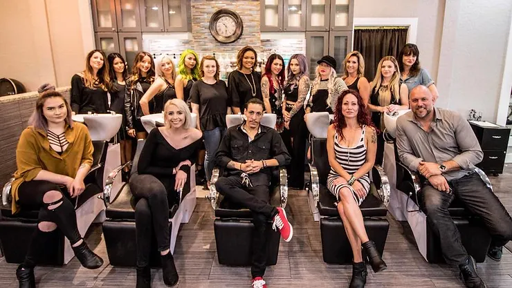 Group of people sitting in a modern salon with bright lighting and contemporary decor. they are posed for a group photo, smiling at the camera, with styling chairs and mirrors in the background.