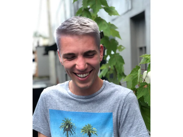 A young man with short gray hair, wearing a gray t-shirt with a palm tree design, laughing with his eyes closed in a blurred green outdoor setting.