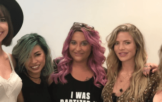 Four women smiling at the camera, each with distinctive hairstyles and colorful hair, ranging from green to pink. they are stylishly dressed and standing close together in an indoor setting.