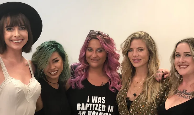 Five women are smiling for a photo. two of them have vibrant hair colors—green and pink. they are fashionably dressed, and one wears a black hat.