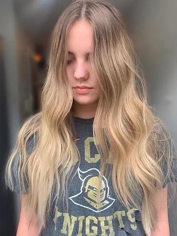 A young woman with long, wavy blonde hair, wearing a dark graphic t-shirt, standing indoors with her eyes closed and a serene expression.