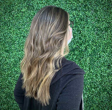 A woman with long, wavy brown hair wearing glasses and a black top, seen from behind against a lush green ivy wall.