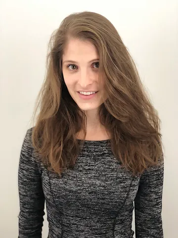 A woman with long, wavy brown hair smiling gently at the camera. she is wearing a gray, textured top and standing in front of a plain white background.