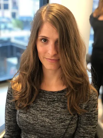 A woman with long brown hair and a subtle smile, wearing a grey knitted top, sitting indoors with a blurred office setting visible through a window in the background.