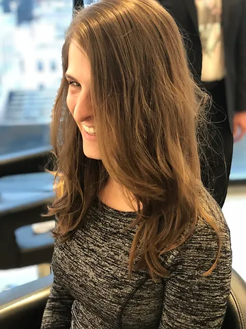 A young woman with long brown hair, wearing a gray knitted top, smiling and glancing to the side in an indoor, well-lit setting.