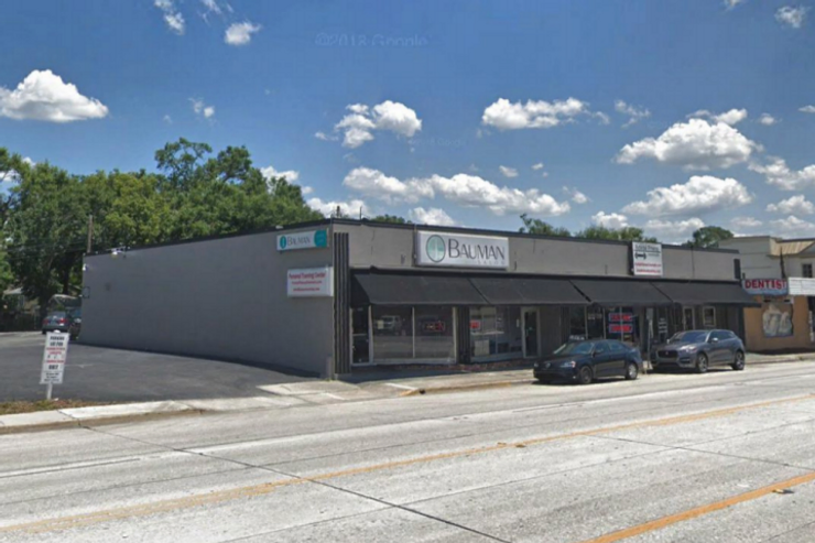 A street view image of a one-story corner building with retail shops, including a sign for "bauman" and another for "dentist," under clear skies with scattered clouds and parked cars.