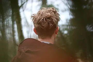 A person with curly blond hair, viewed from behind, wearing a brown jacket and standing in a forest with blurred trees in the background.