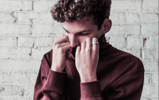 A young man with curly hair, wearing a maroon sweater and a silver ring, appears contemplative as he holds his nose with both hands against a white brick background.