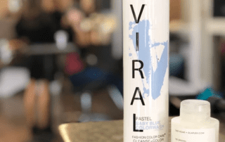 Close-up of a vira pastel baby blue colorwash bottle in a salon, with a smaller olaplex bottle in the foreground and blurry figures in the background.