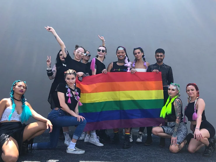 A diverse group of people smiling and posing with a pride flag at an outdoor event, expressing joy and unity. they are dressed in colorful and vibrant outfits, featuring dyed hair and sunglasses.