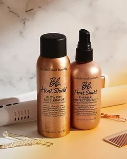 Two bumble and bumble styling products, heat shield blow dry accelerator and thermal protection mist, on a counter with a marble backdrop, next to hairpins and a hairdryer.